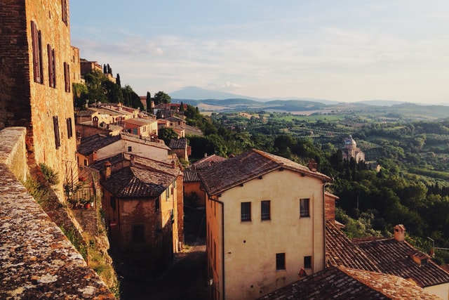 Orange-brown plaster and brick buildings cascade down a hillside; in the distance a church dome, and beyond it rolling hills fade into the Tuscan haze